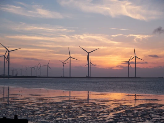 Turbina wiatrowa stoi wysoko na farmie wiatrowej nad wodą o zmierzchu, tworząc piękne światło popołudniowe na horyzoncie. Niebo jest pomalowane płynnymi odcieniami, pokazując znaczenie zasobów wodnych w energii odnawialnej.